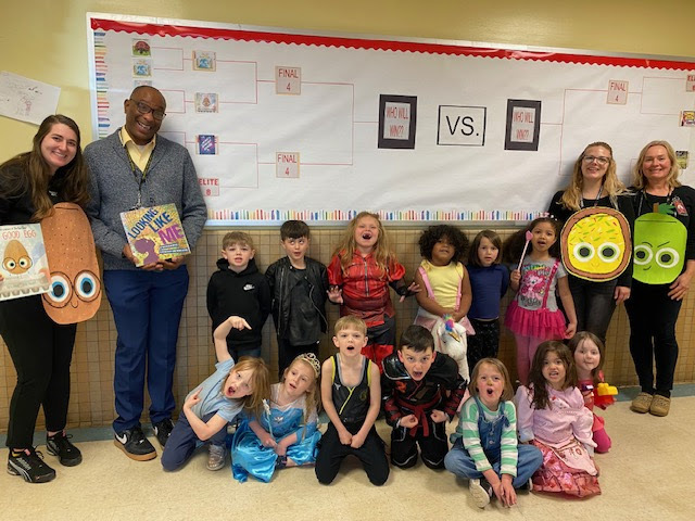 A group of elementary school students and four adults stand in front of a bulletin board decorated with a book-themed tournament bracket. The children are dressed in costumes, including princess dresses and superhero outfits, while two of the adults wear costumes shaped like book characters and one holds a picture book. Everyone is smiling and posing together in a cheerful classroom setting.