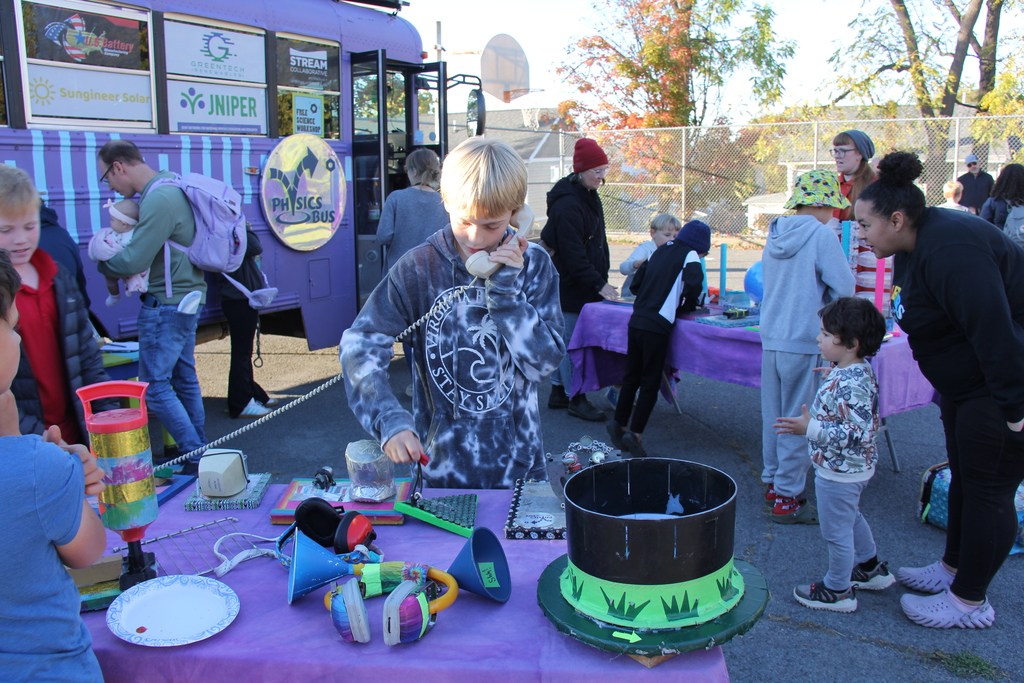 Children and adults explore science exhibits outside the purple Physics Bus. Tables covered with purple cloth display colorful hands-on experiments and recycled materials. A boy speaks into a retro phone while others look on.