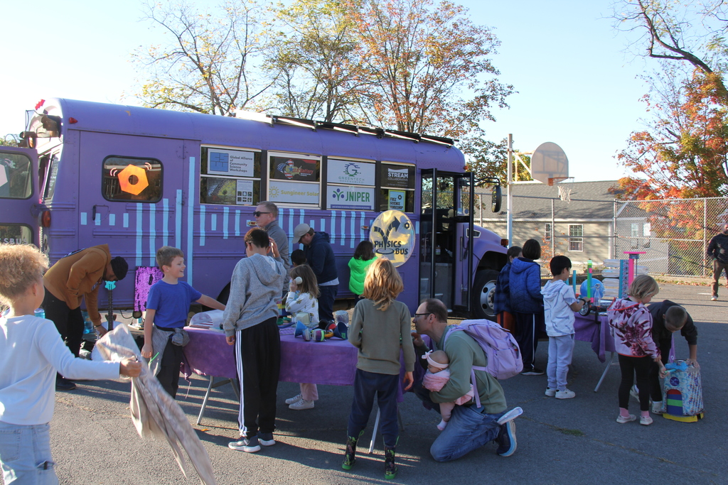 Children and adults gather around a purple “Physics Bus” parked on a playground. Tables are set up with hands-on science activities, and the scene is bright and sunny with fall foliage in the background.