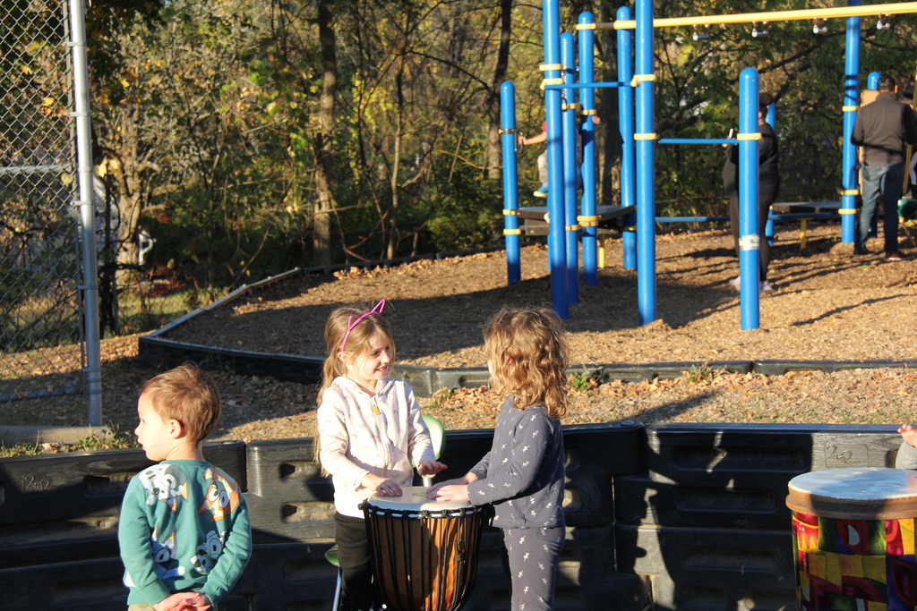 Three young children play outdoors near a playground. Two girls are smiling and drumming together on a hand drum while a boy stands nearby. Blue and yellow climbing equipment is visible in the background.
