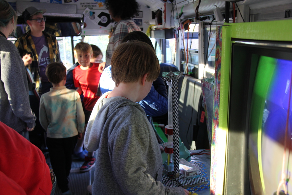 Students explore a science exhibit inside a brightly decorated bus filled with interactive displays and equipment, illuminated by sunlight streaming through the windows.