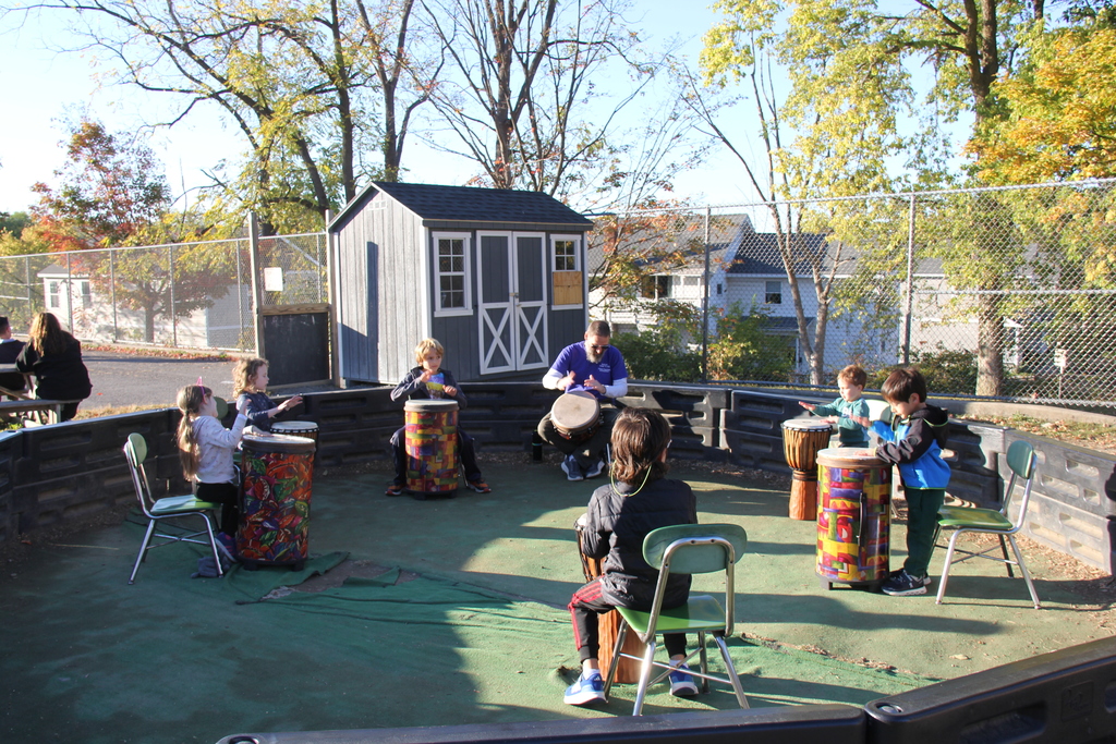 A group of children sit and stand in a semicircle outdoors, playing drums of various sizes with an instructor. The drumming circle takes place on a fenced playground area surrounded by trees with autumn leaves.A group of children sit and stand in a semicircle outdoors, playing drums of various sizes with an instructor. The drumming circle takes place on a fenced playground area surrounded by trees with autumn leaves.