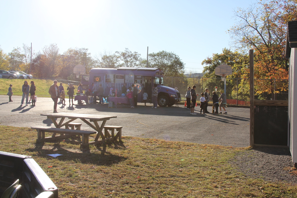 Families and children explore the purple “Physics Bus” and nearby activity tables on a sunny afternoon. The scene is viewed from a distance, with picnic tables and trees with colorful fall leaves in the foreground.