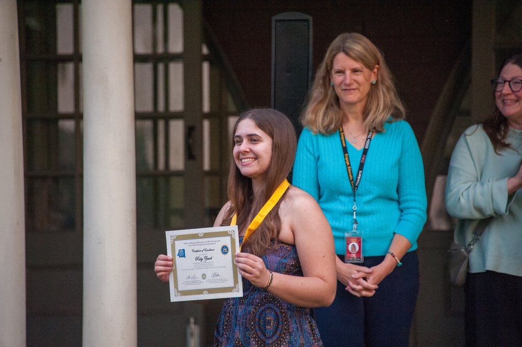 A smiling student stands outdoors holding a certificate and wearing a yellow honor cord around their neck. Two adults stand behind them, also smiling, during what appears to be an award or recognition ceremony.