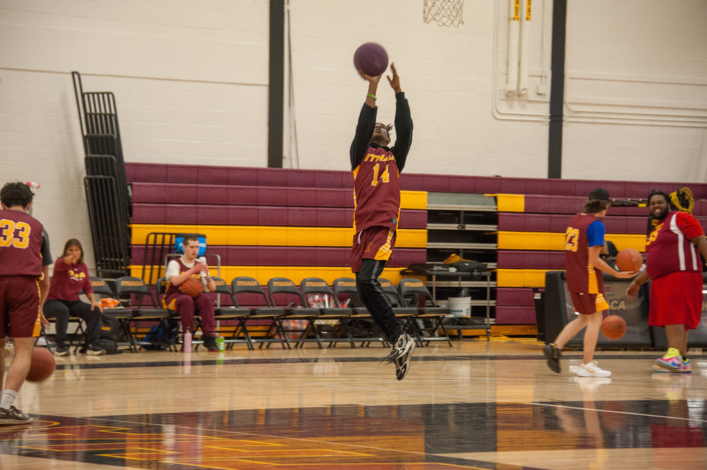 A basketball player wearing a maroon and gold uniform with the number 14 jumps toward the hoop for a layup during a game in a gymnasium. Other players and coaches are visible in the background near the benches.