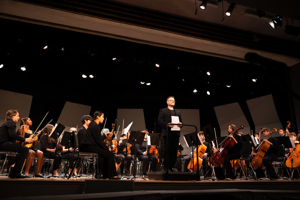 A conductor stands on a podium addressing the audience while holding sheet music, surrounded by a seated student orchestra on stage. The musicians, dressed in black, hold violins, cellos, and other instruments under bright stage lights.