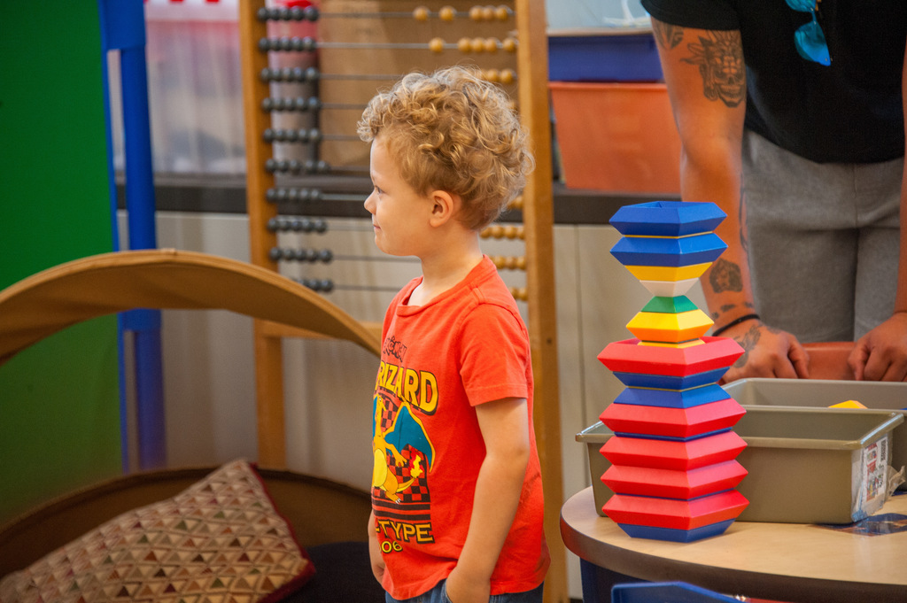 A small child in a red shirt stands and looks off into the classroom. He is standing beside blocks.