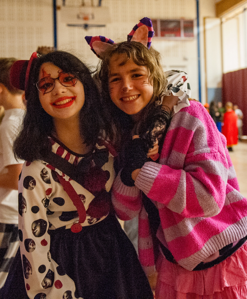 Two smiling students in Halloween costumes pose together in a school gym. One wears a black-and-white polka-dot outfit with red suspenders and face paint, while the other wears pink-and-purple striped cat ears and a matching sweater. They stand close, enjoying the celebration.