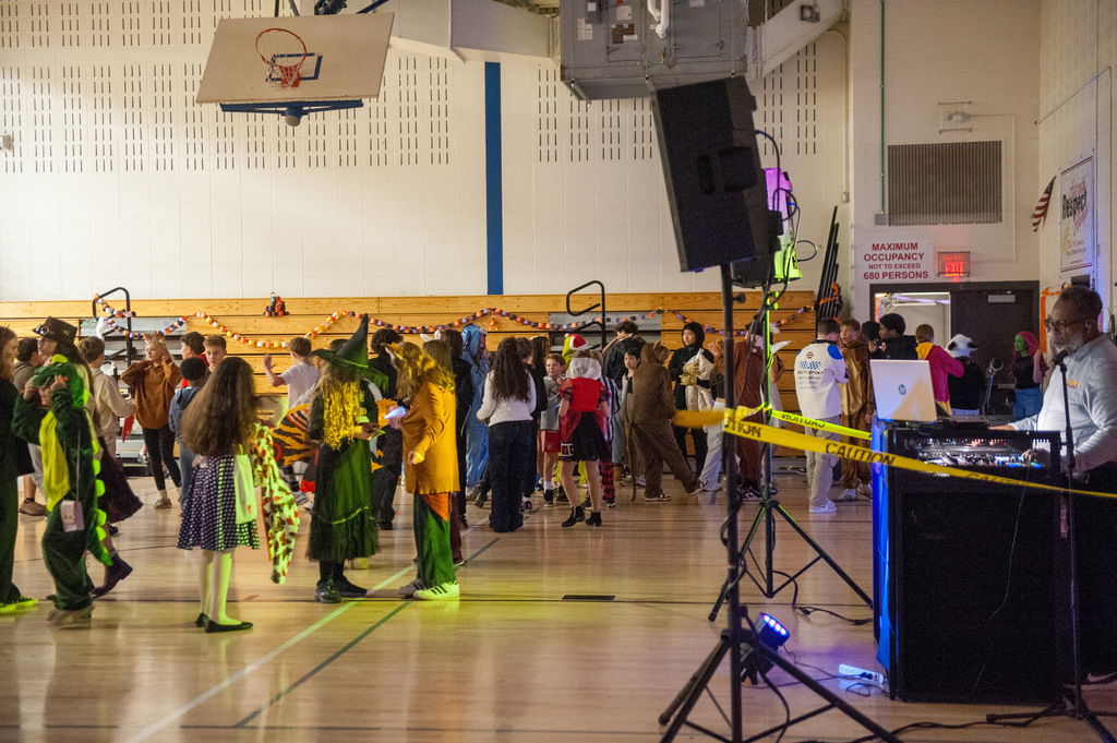 A wide shot of a school gym filled with students in Halloween costumes dancing and socializing. Decorations like paper chains and caution tape line the space, and a DJ operates sound equipment on the right side of the image. The gym’s basketball hoops and stage lights are visible above.