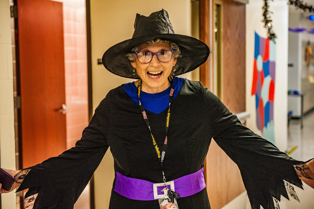 A smiling adult wearing a black witch costume with a wide-brimmed hat and purple belt stands in a school hallway with arms outstretched. Classroom doors and colorful decorations are visible in the background.