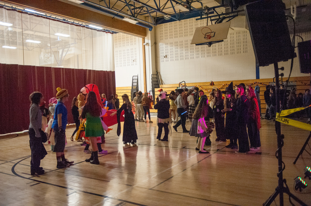Students in a variety of costumes gather and dance in a dimly lit school gym decorated for a Halloween celebration. Bleachers and basketball hoops are visible in the background.