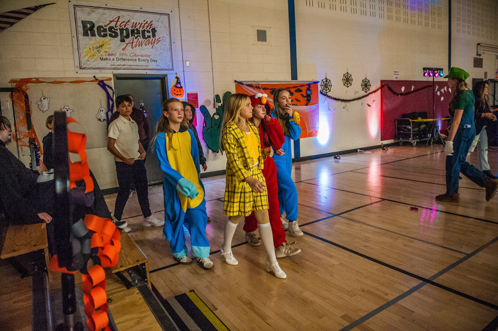 A group of students wearing costumes, including a yellow plaid dress and colorful character onesies, walk together across a decorated school gym floor. Halloween decorations and a sign reading “Act with Respect Always” hang on the wall behind them.