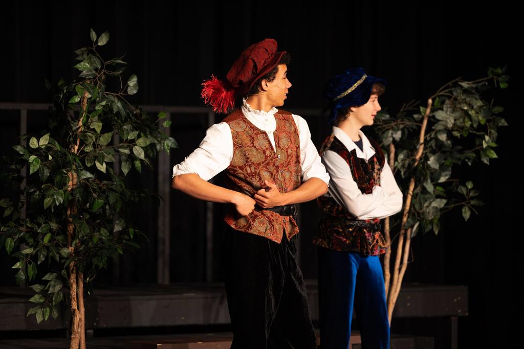 Two young actors on stage dressed in Renaissance-style costumes, including patterned vests and floppy hats with feathers. They stand beside potted trees, looking off to the left as if reacting to something happening offstage.
