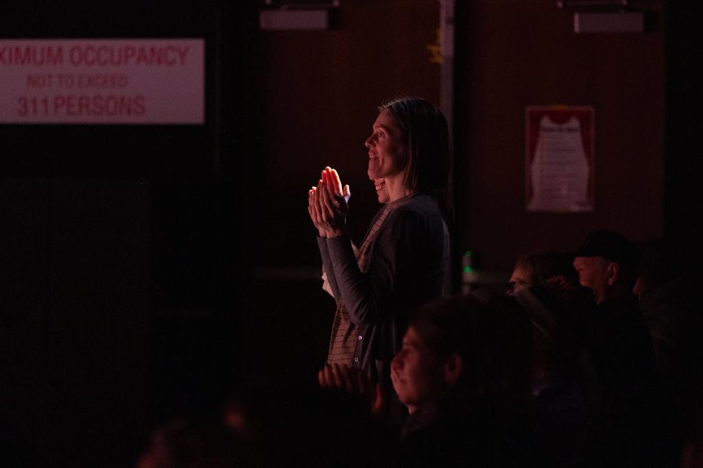 A person standing in a dimly lit auditorium, warmly illuminated by stage light, clapping while watching a performance. Other audience members sit in the shadows around them.