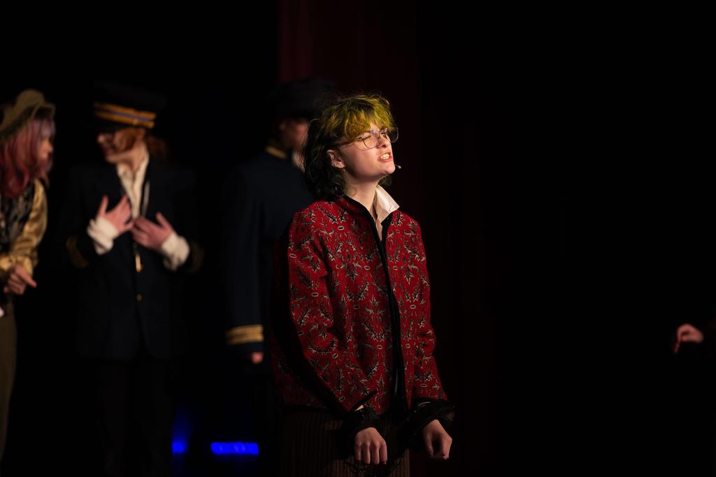 A student in a red patterned jacket, wearing glasses and a stage microphone, stands under a spotlight delivering a theatrical line while other performers in costume stand blurred in the background.