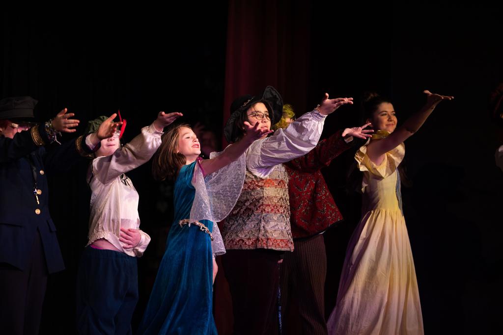 A group of student performers on stage, reaching their arms outward in unison while wearing colorful period-style costumes during a theatrical production.