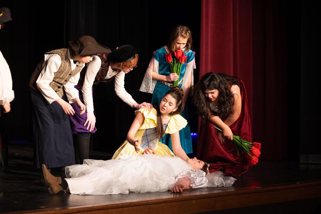 A group of young actors in colorful period costumes gathers around another performer lying on the stage in a white gown, portraying a dramatic or emotional moment. Several students kneel or lean in, one holding red flowers, as they react with concern.