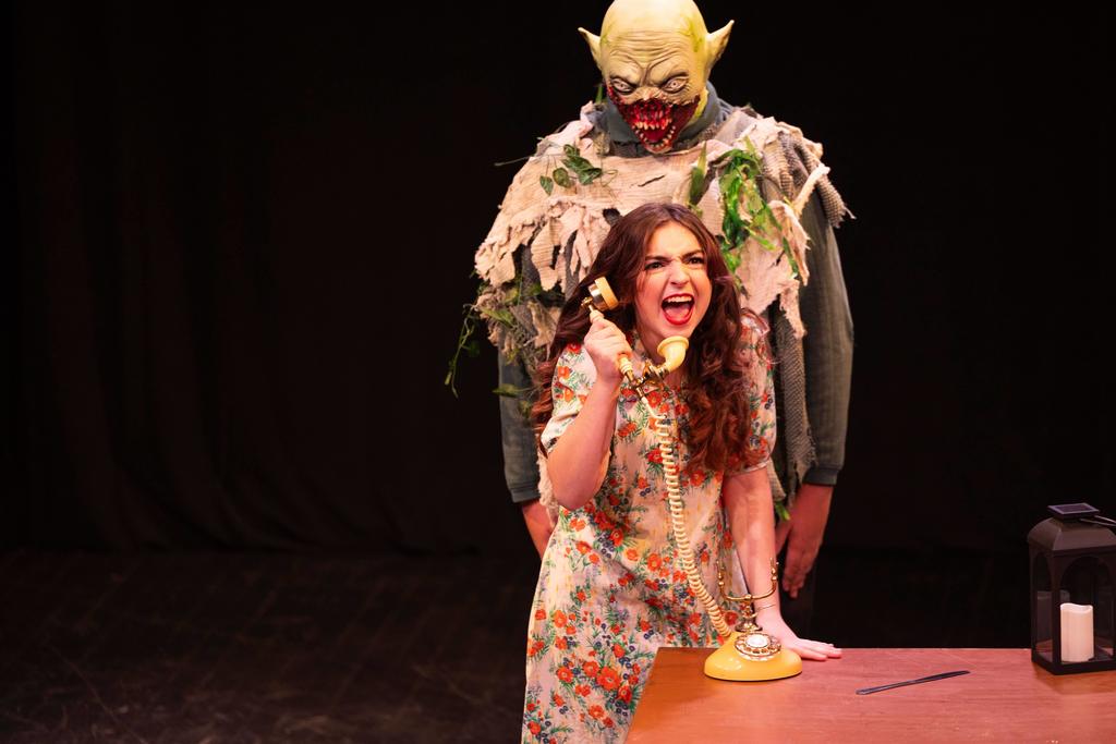 A stage performance scene shows a young woman in a floral dress leaning over a desk, angrily shouting into a yellow rotary telephone. Behind her stands a tall, ominous figure in a tattered costume with a grotesque mask, looming closely. The background is dark, with stage lighting focused on the performers and a desk with props in the foreground.