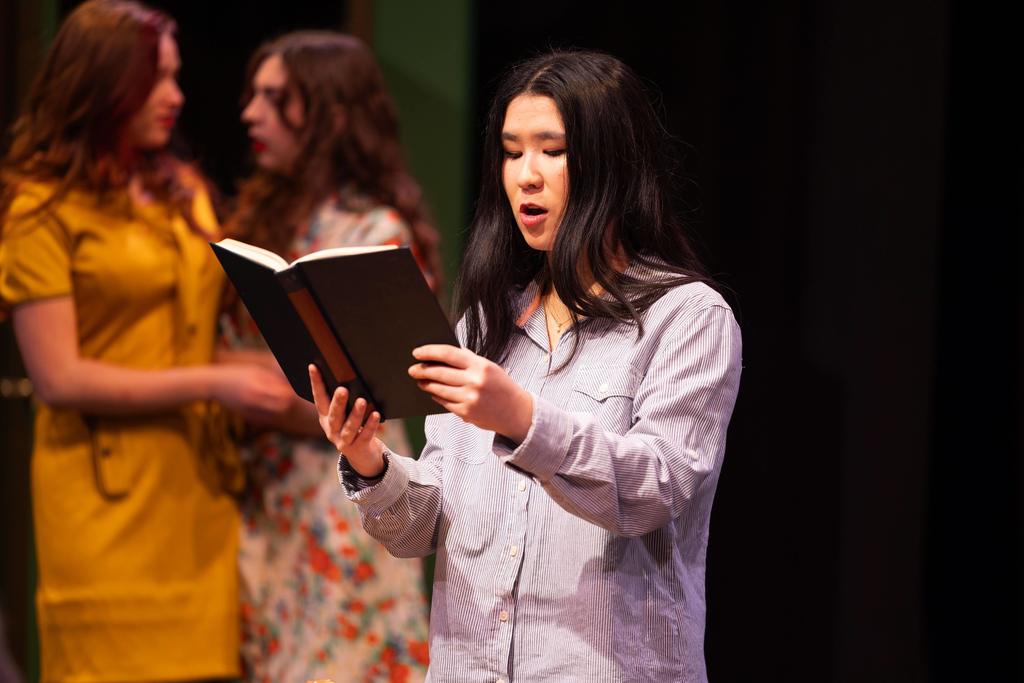 A student stands on a stage reading aloud from a black book, focused and mid-speech. Behind them, two other students in dresses are slightly out of focus, suggesting an ongoing scene in a theatrical performance under stage lighting.