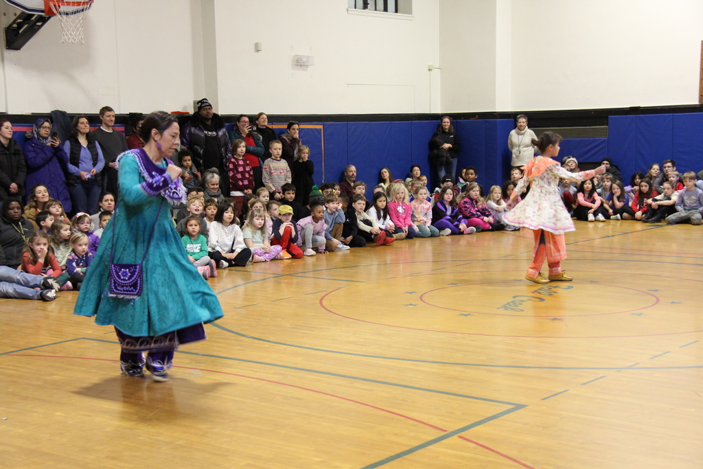 Two performers in bright, traditional outfits dance at the center of a school gym floor while rows of young students sit cross-legged along the wall, watching attentively. Families and staff stand behind the seated children.