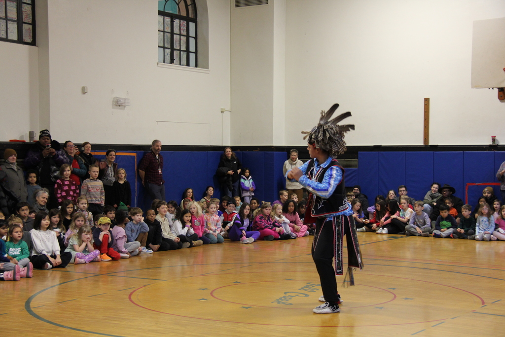 A visiting performer wearing a feathered headdress and traditional attire stands at the center of a school gym, gesturing as they address a large group of seated elementary students. Adults stand along the gym walls observing.