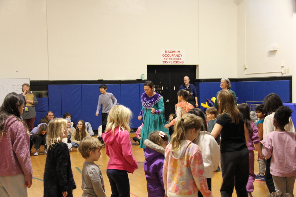 Children and adults stand and move together on a school gym floor as a performer in a teal and purple traditional dress leads an interactive activity. Students of various ages participate while others sit or watch nearby.