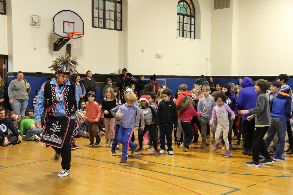 A performer wearing a feathered headdress leads a group of elementary students in a dance across a gym floor. Children follow closely behind, smiling and moving together, as adults observe from the edges of the room.