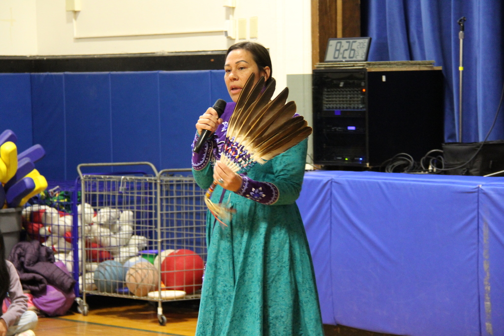 A performer in a teal traditional dress speaks into a microphone while holding a feather fan in a school gym. Audio equipment and gym supplies are visible behind her as she addresses the audience.