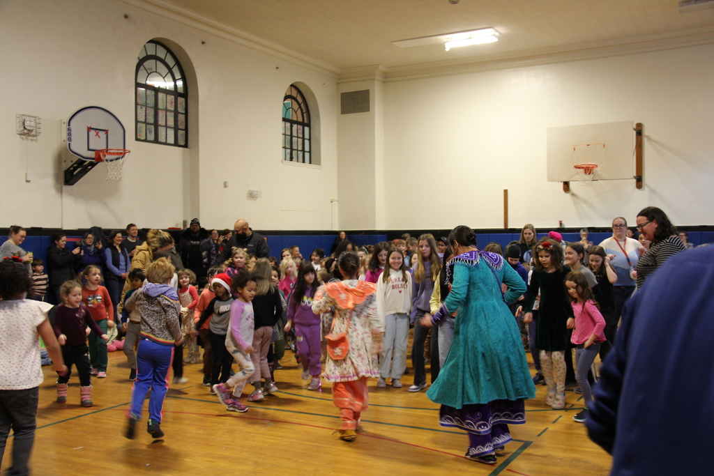 A large group of elementary-aged children dance together in a school gymnasium while adults watch from the sidelines. Two visiting performers in colorful traditional clothing move among the students, encouraging participation.