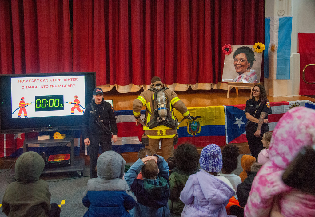 A firefighter demonstrates putting on full protective gear, including an air tank, on a school auditorium stage while children watch from the floor and other firefighters stand nearby.