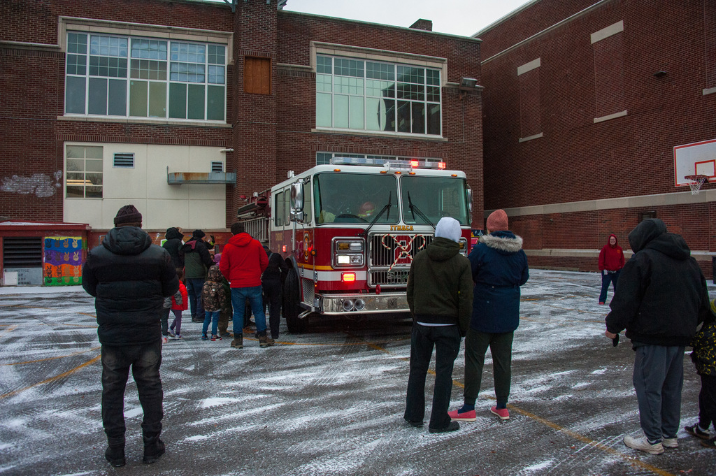 A fire truck is parked in a snowy schoolyard outside a brick school building while children and adults in winter coats stand nearby, watching and gathering around the truck.