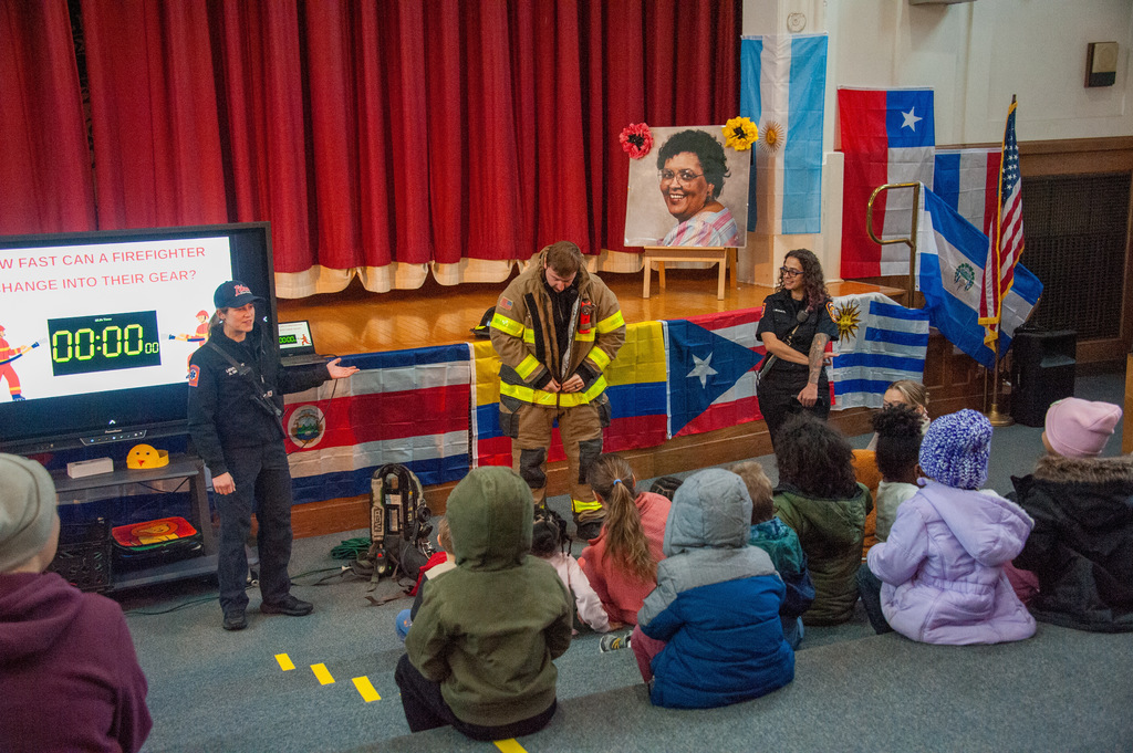In a school auditorium, firefighters demonstrate protective gear on stage while young children sit on the floor watching. A screen displays a timer for how fast a firefighter can change into gear, with international flags hanging behind the stage.