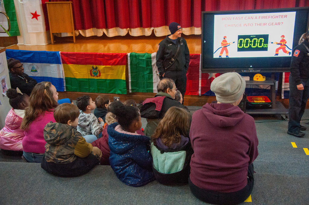 Children and adults sit on the floor of a school auditorium facing firefighters, who are standing near a large screen displaying a timer and the question, “How fast can a firefighter change into their gear?”