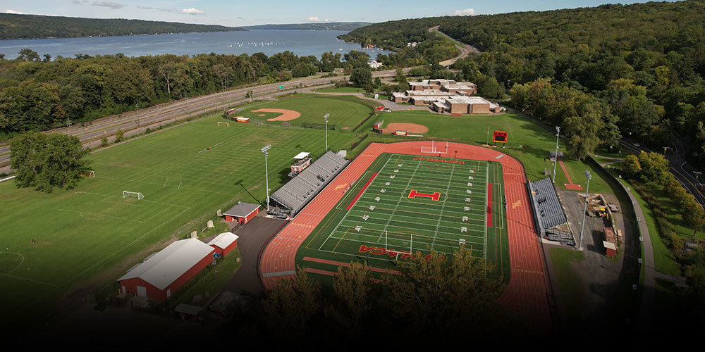 Aerial view of a IHS athletic complex with a football field and red running track at center, bleachers on both sides, adjacent soccer fields and a baseball diamond, surrounded by trees and hills, with a large lake visible in the background.