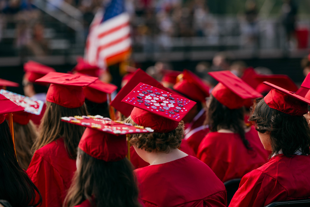 IHS graduates wearing red caps and gowns sit together at a commencement ceremony, with decorated mortarboards visible and a blurred American flag and audience in the background.