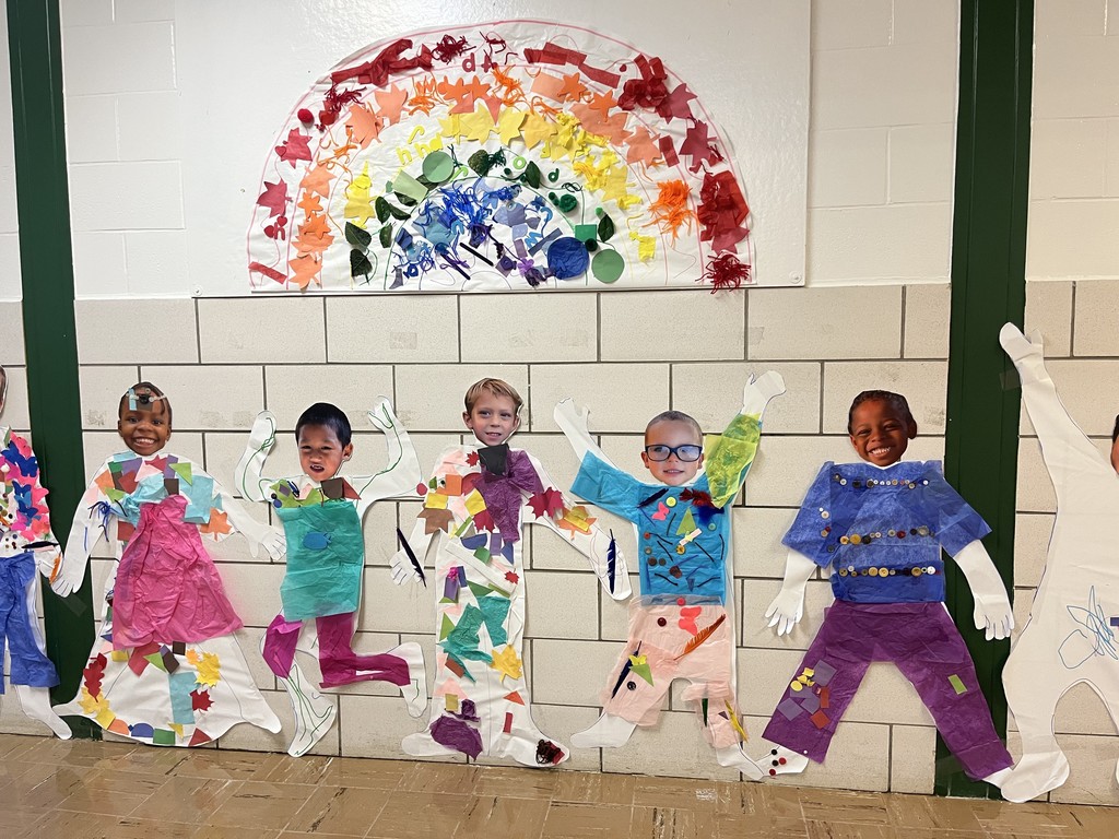 A hallway display shows five smiling elementary-aged children standing in front of a tiled wall, each with their face placed on a life-size paper cutout body decorated with colorful tissue paper, buttons, and craft materials. The figures pose with raised arms and playful stances, creating a sense of movement. Above them hangs a large, handmade rainbow collage made from layered paper, leaves, and textured materials in red, orange, yellow, green, blue, and purple.