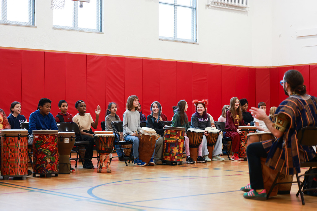 Elementary students sit in a semicircle playing West African drums as an instructor leads the group during a cultural music workshop in the gym.