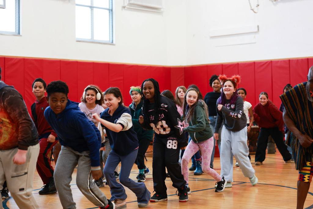 Elementary students smile and move together while learning West African dance in a school gymnasium with red padded walls.