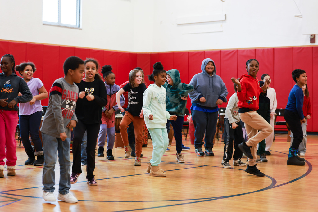 A diverse group of elementary-age children dance together in a school gymnasium with a wooden floor and red padded walls. The kids are mid-movement, smiling and stepping in rhythm, wearing casual winter clothes like hoodies, sweatpants, and sneakers.
