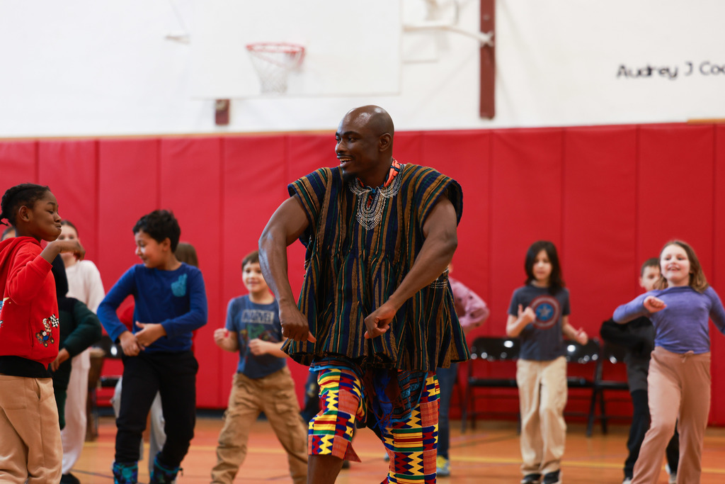 A visiting artist demonstrates West African dance movements while elementary students follow along in a gymnasium.