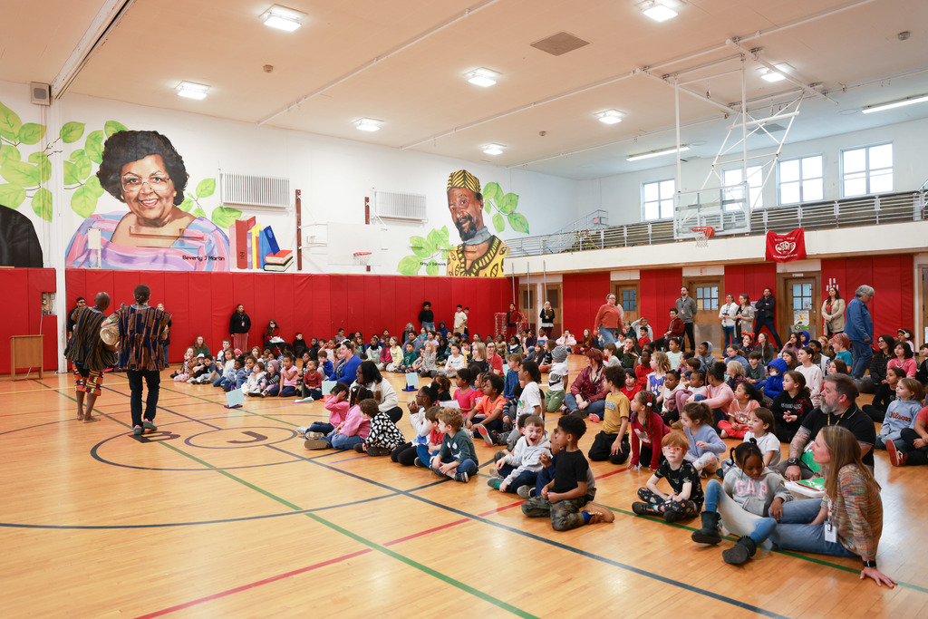 A large group of children sit on the floor of a school gymnasium facing two performers standing at the front. The gym has red padded walls and colorful murals of people and books. Teachers and adults stand along the walls while the children watch attentively, suggesting a school assembly or cultural performance.