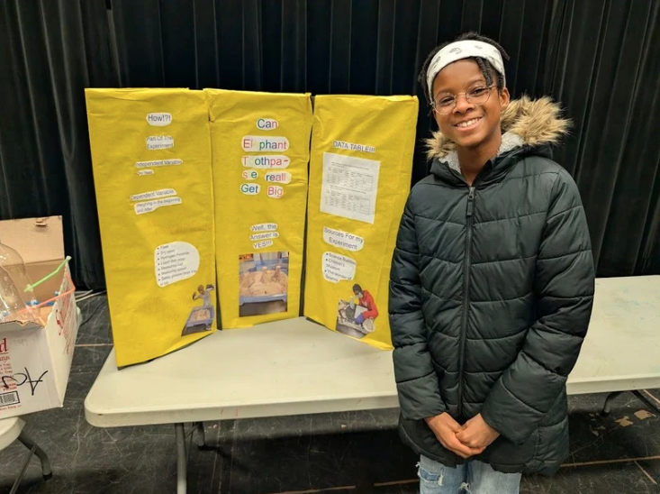 An LACS 6th-grader stands in front of their yellow poster board at the science fair
