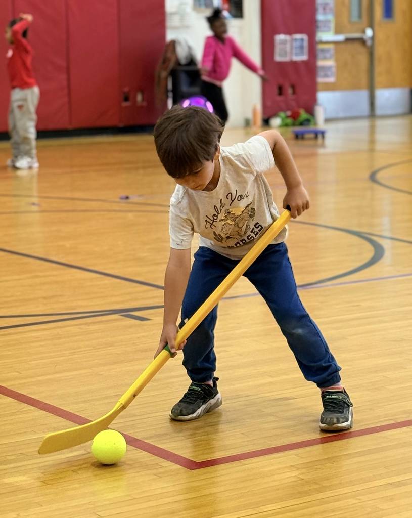 A BJM student in the gym holding a hockey stick, setting up to hit a ball.