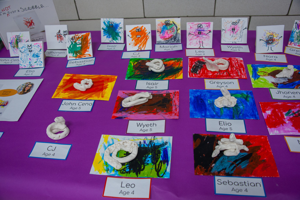 Colorful artworks, including painted cards and clay figures, displayed on a purple table. Each piece has a label with names and ages.