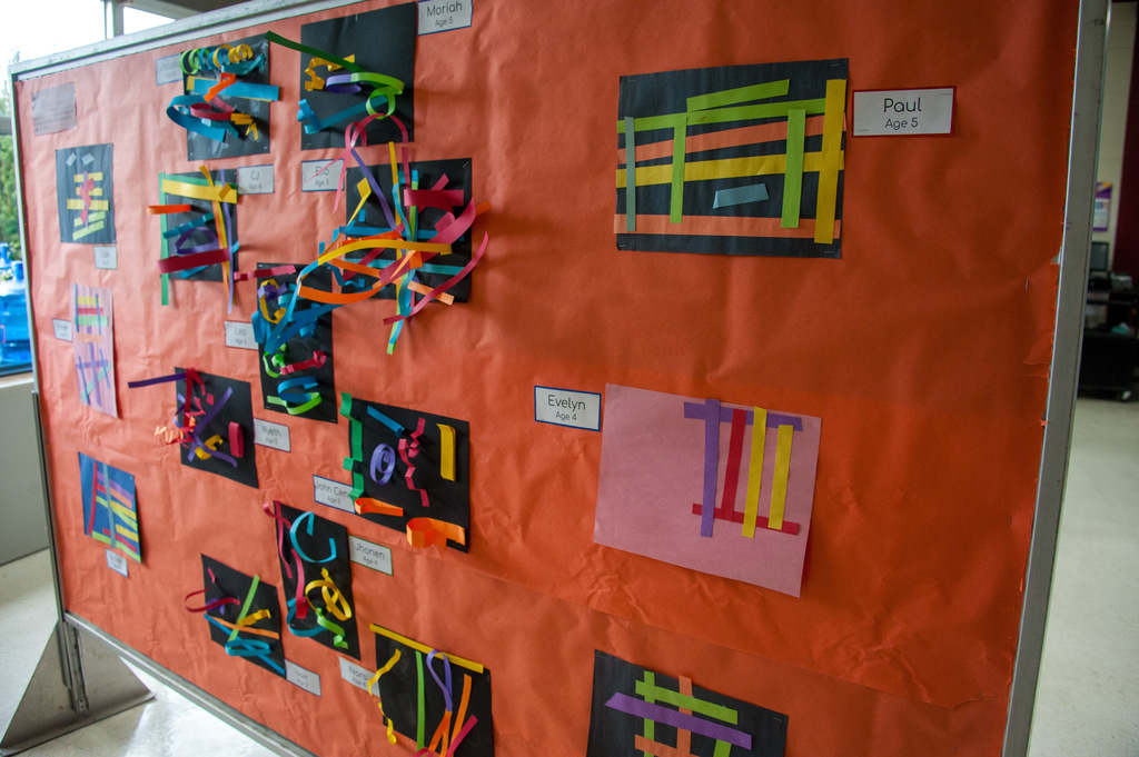 An orange bulletin board displays various colorful artwork on black backgrounds, with each piece labeled. The board is located in an indoor space.