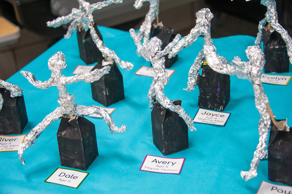 Figures made of aluminum foil attached to black bases with name tags. Displayed on a blue table.