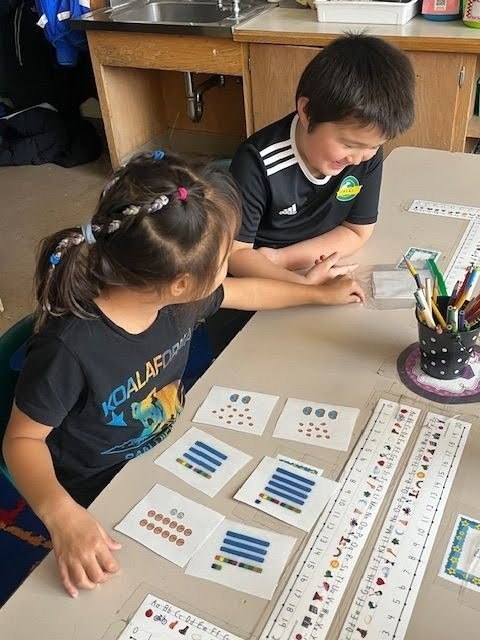 Two children at a table with educational materials, one leaning over the table and the other looking at cards.