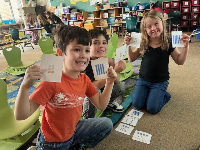 Three kids sit on the floor in a classroom. They hold up cards with numbers and smile.