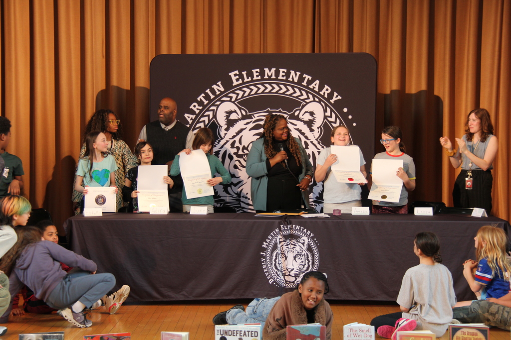 A group of children and adults hold certificates while seated and standing on a stage, with a tiger logo behind them.