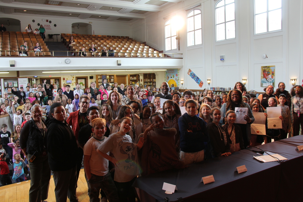Large group of people in a hall with rows of empty seats. Some hold signs. A long table with items in front.